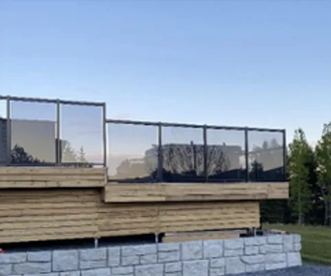 Wooden deck with a glass railing sitting on a stone-block base against a blue evening sky