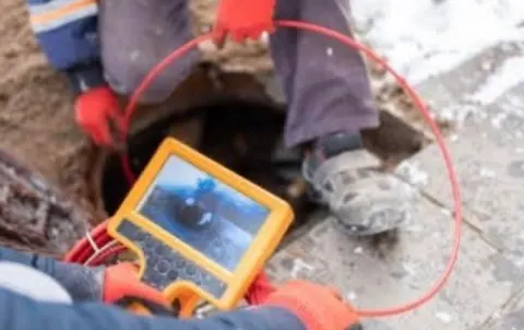 Worker feeding a red camera cable into a sewer access hole while watching a handheld yellow monitor