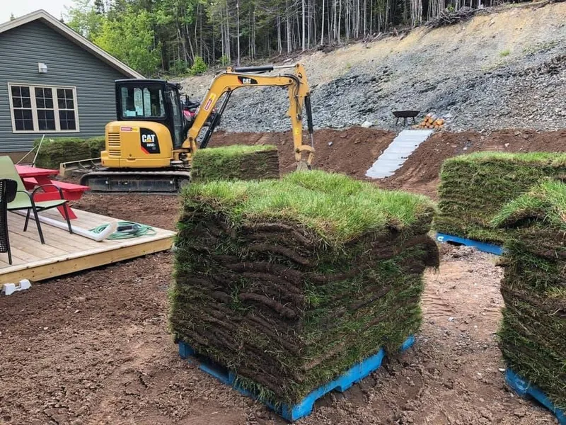 Pallets of sod beside a new wooden deck with a yellow excavator regrading a slope behind them